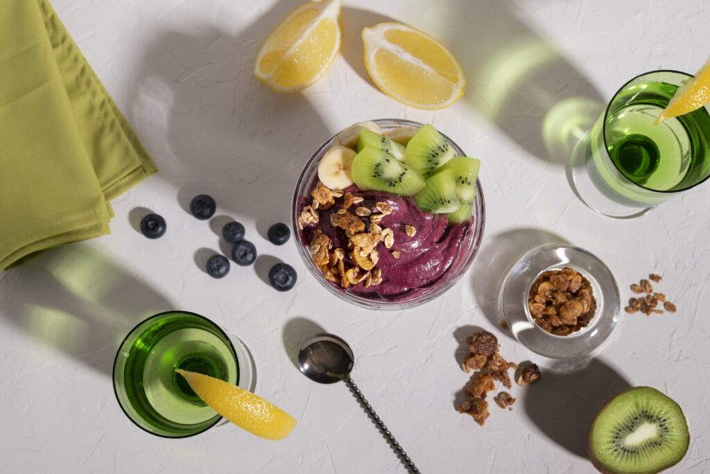 Imagine de sus cu un pahar de smoothie cu kiwi pentru slabit asezat pe o masa alba, inconjurat de alte ingrediente: afine, lamai, semințe