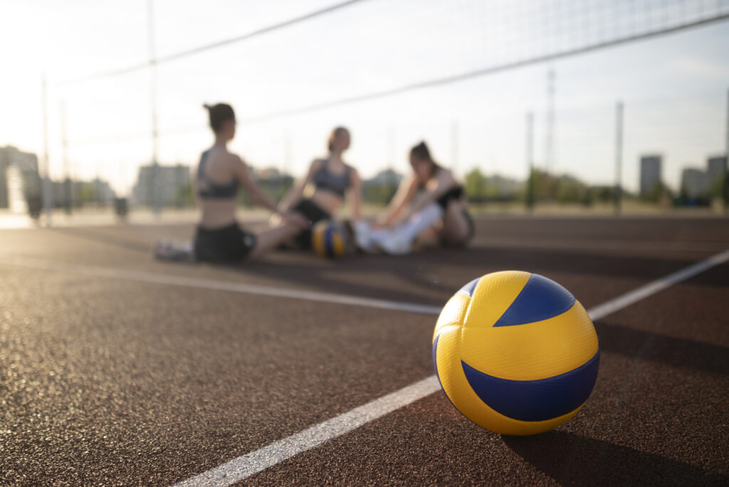 Imagine de aproape cu minge de volei pe un teren sport, in spate un grup de fete