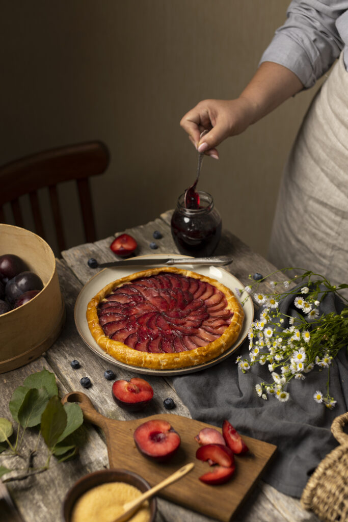 Tarta cu prune pe o masa cu decor rustic. In colt, mana unei persoane ia o lingura de gem cu prune ca sa decoreze tarta fara zahar