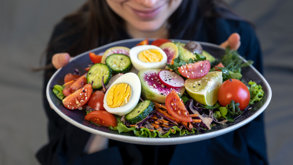 Imagine de aproape cu o farfurie plină de alimente proaspete, sănătoase: ouă, roșii, smochine, lime, într-o salată. Farfuria e ținută în mâini de o femeie, care se vede zâmbind în fundal