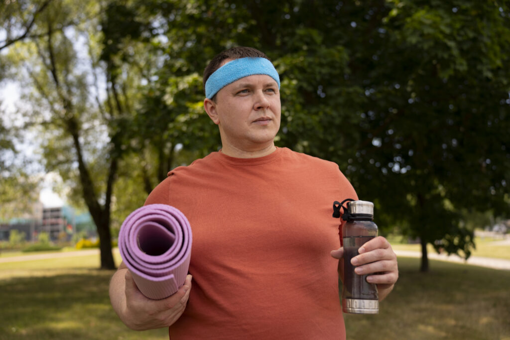 Bărbat îmbrăcat în echipament sport. Într-o mână ține o sticlă de apă, iar în cealaltă un covoraș de antrenament. Imagine afară