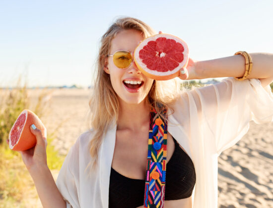 Fată tânără blondă ține 2 bucăți de grepfrut și le arată fericită către cameră
