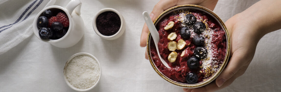Imagine de sus cu mâinile unei persoane care ține un bol cu desert de dietă: smoothie bowl cu iaurt, fructe de pădure, fulgi de cocos