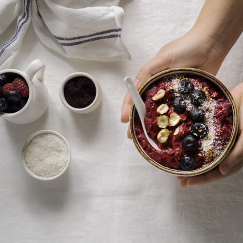 Imagine de sus cu mâinile unei persoane care ține un bol cu desert de dietă: smoothie bowl cu iaurt, fructe de pădure, fulgi de cocos