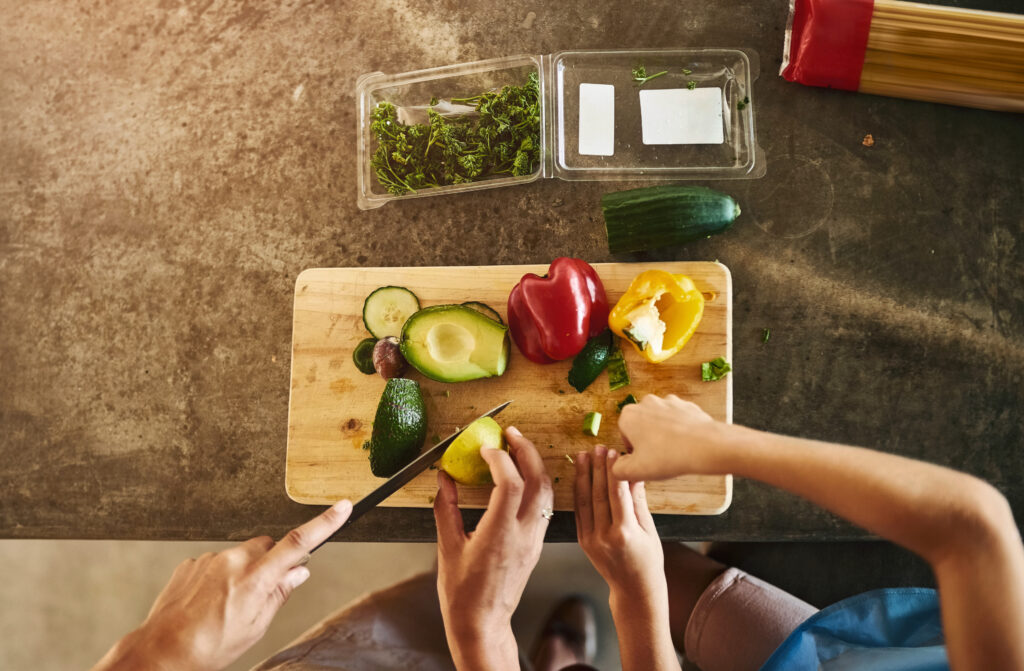 Imagine de sus, persoane care taie legume proapete pentru prepararea mesei: avocado, ardei gras, saladă, dovlecel verde, alimente permise și încurajate în dieta Dean Ornish