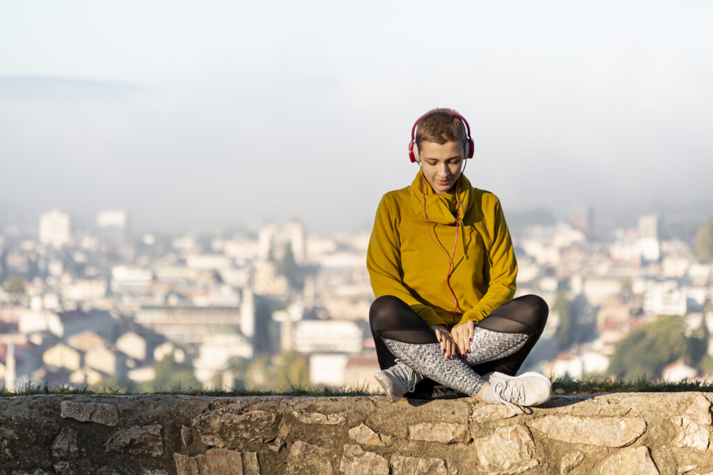 Fată tânără ascultă muzică la căști în timp ce stă pe un zid de piatră. În spate, imagine panoramică a orasșului