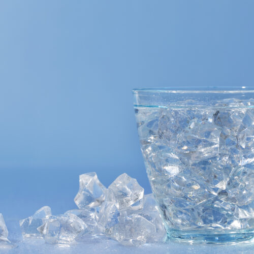 un pahar de apa plin cu cuburi de gheata, cuburi de gheata pe langa pahar, fundal albastru