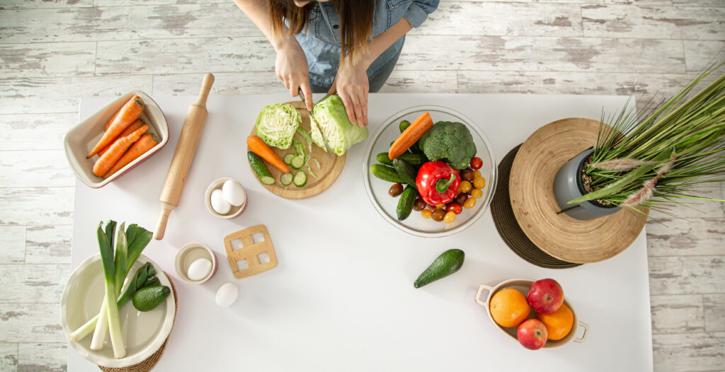 Fewmeie tânără prepară o salată sănătoasă în bucătărie. Imagine de sus. Pe masă, sunt diverse fructe și legume, ouă și un făcăleț de lemn