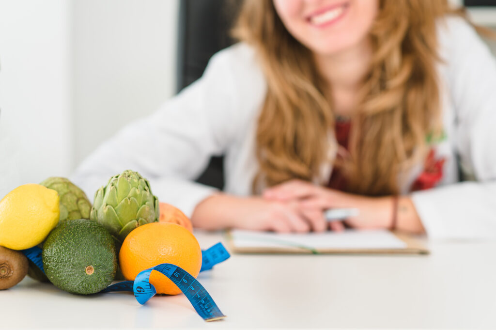 Imagine de aproape cu fructe sănătoase, precum avocado, portocale, kiwi și lămâi, așezate pe o masă. Ele sunt înconjurate de un centimetru albastru. Imaginea este din cabinetul unui nutriționist, care stă în fundal