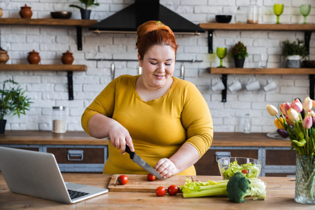 Imagine din față, persoană supraponderală care gătește un preparat sănătos din dieta Cambridge. Urmărește pașii de preparare de pe laptop