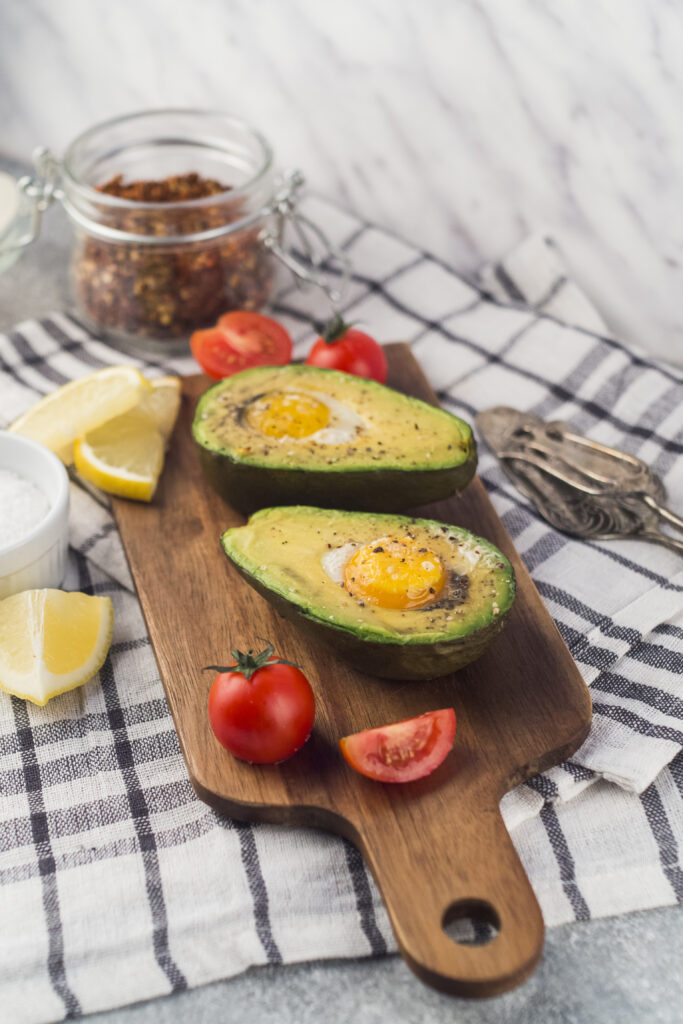 Două jumătăți de avcocado copt cu câte un ou ochi fiecare. Bucățile de avocado sunt așezate pe un tocător de lemn, înconjurat de roșii cherry, lămâi, condimente.