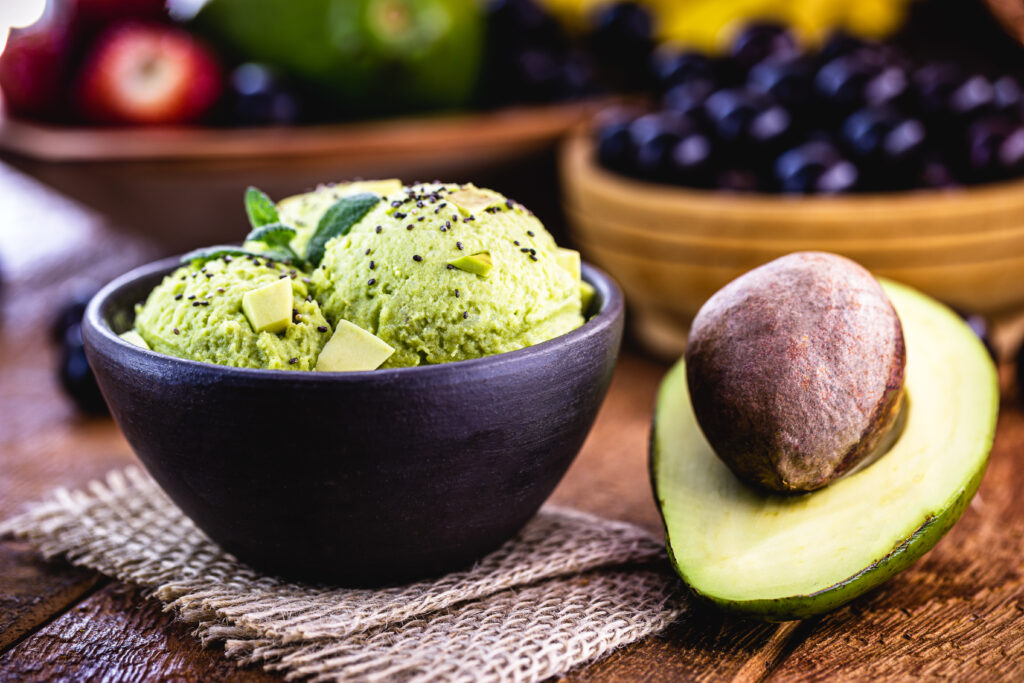 Înghețată de avocado, în bol negru. Lânmgă, o jumătate de avocado. Multe alte fructe pot fi văzute în spate, în fundal