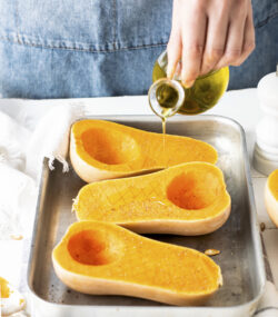 bucati de dovleac taiate pe jumatate pe tava, mana unei femei adauga peste ulei de măsline