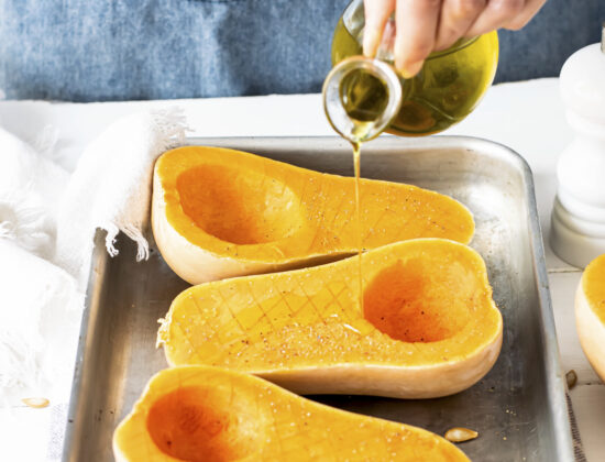 bucati de dovleac taiate pe jumatate pe tava, mana unei femei adauga peste ulei de măsline