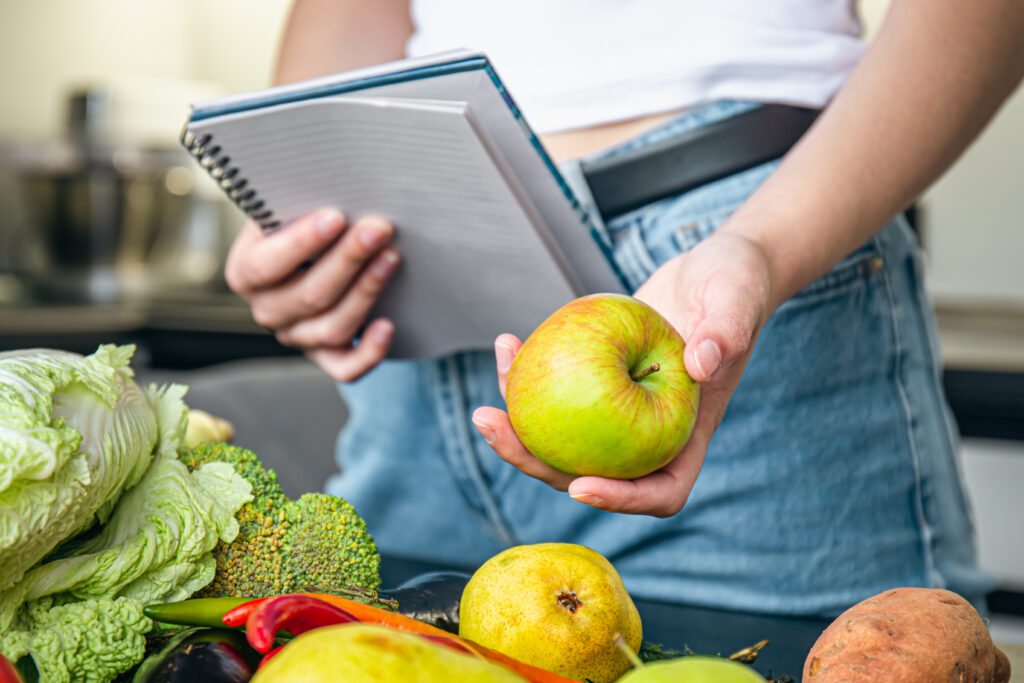 Imagine de aproape, femeie care ține într-o mână un măr și în cealaltă un carnețel pentru a calcula deficitul caloric. Pe masă sunt și alte legume proaspete, potrivite într-un meniu pentru dieta alcalină