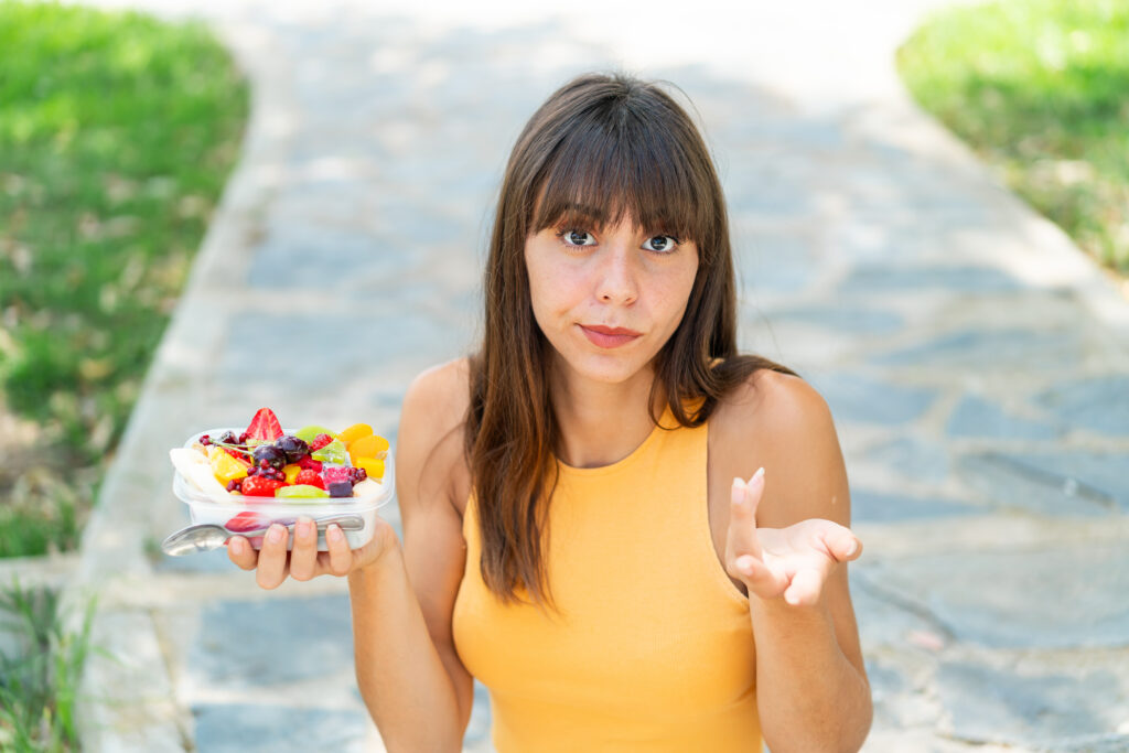 Femeie tănără, se uită întrebătoare către cameră. Ține în una dintre mâini o caserolă cu fructe proaspete, alimente permise în dieta de 800 calorii.