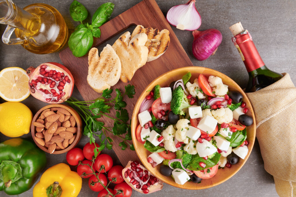 Preparat în dieta zonelor albastre: salată grecească cu legume, brânză și rodie, într-un bol așezat pe un tocător de lemn. În jur, numeroase alte fructe, legume, migdale. Lângă, o sticlă de vin