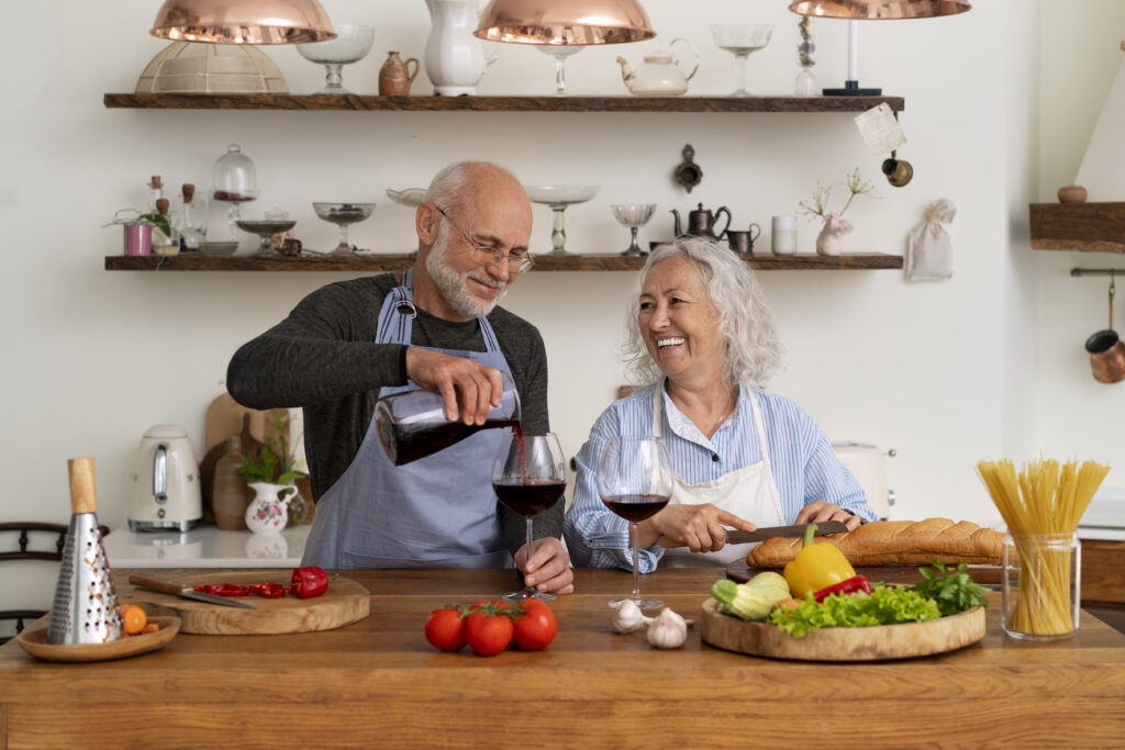Cuplu vârstnic la un pahar de vin, în bucătărie, în timp ce pregătesc un preparat sănătos, specific în dieta zonelor albastre