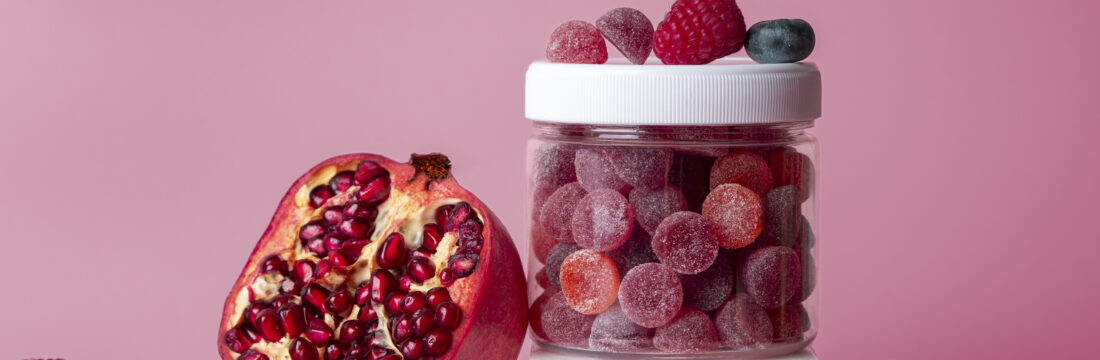 Borcănel de suplimente alimentare gummies roșii și mov, cu resveratrol. Lângă, o rodie, aliment care conține resveratrol. Fundal roz în spate