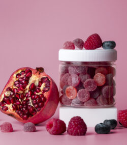 Borcănel de suplimente alimentare gummies roșii și mov, cu resveratrol. Lângă, o rodie, aliment care conține resveratrol. Fundal roz în spate