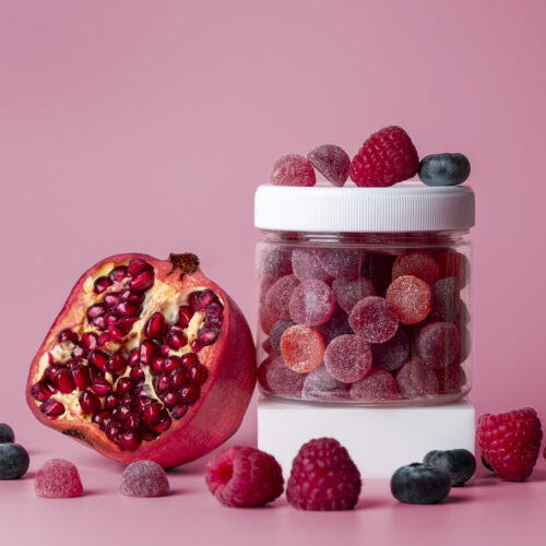 Borcănel de suplimente alimentare gummies roșii și mov, cu resveratrol. Lângă, o rodie, aliment care conține resveratrol. Fundal roz în spate