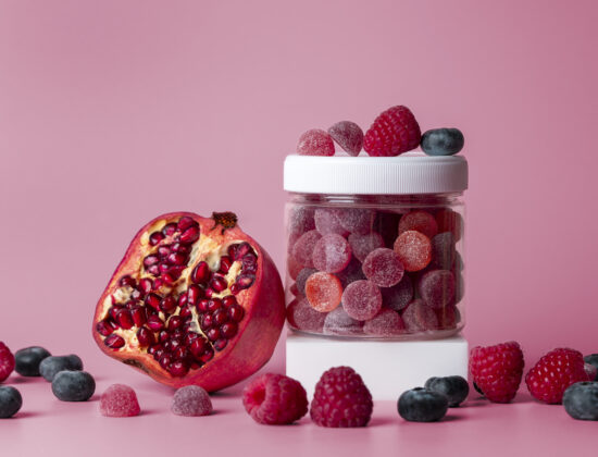 Borcănel de suplimente alimentare gummies roșii și mov, cu resveratrol. Lângă, o rodie, aliment care conține resveratrol. Fundal roz în spate