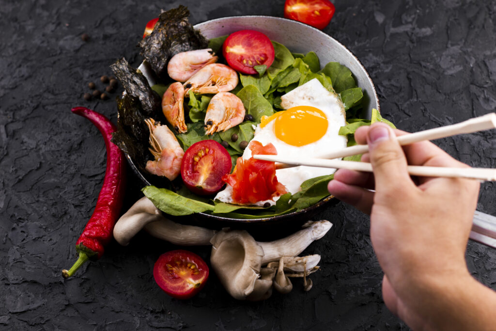 Imagine de sus, persoană care mănâncă dintr- o salată cu ou ochi și creveți, plus spanac și legume. Se vede doar mâna persoanei, care ia din legume cu bețișoare de mâncat