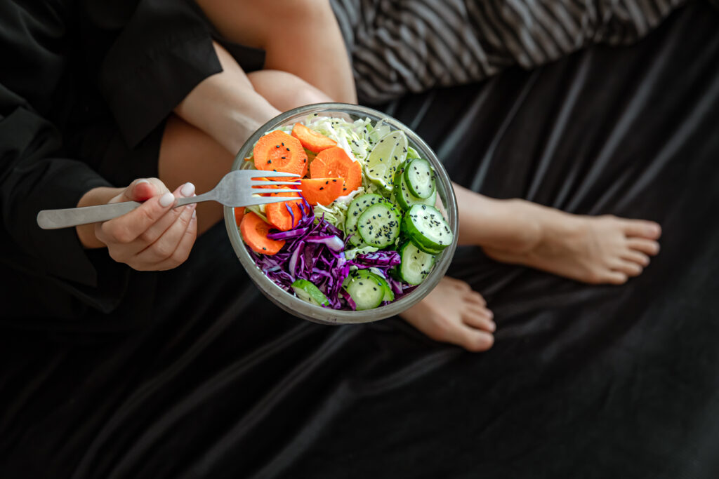 Imagine de sus, persoană care stă în pat și mănâncă un bol de salată. În fundal i se văd doar picioarele
