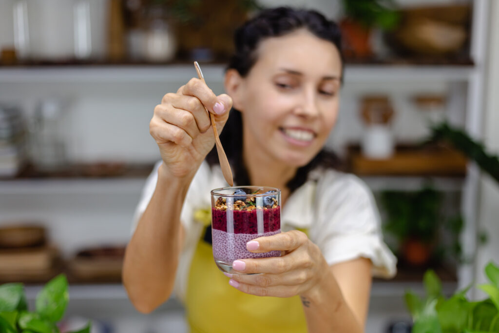 Femeie care arată către cameră un borcănel transparent cu budincă de chia cu fructe de pădure. Imagine din față, femeia ține în mână o lingură de lemn pe care se pregătește să o bage în borcănel