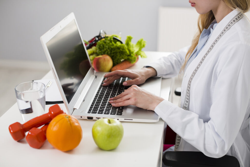Nutriționist cercetează în fața unui laptop. Imagine de aproape. Pe birou: fructe, un pahar cu apă, un stetoscop, echipament sport