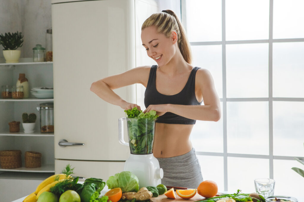 Femeie tânără îmbrăcată în echipament sport își pregătește un smoothie cu verdețuri