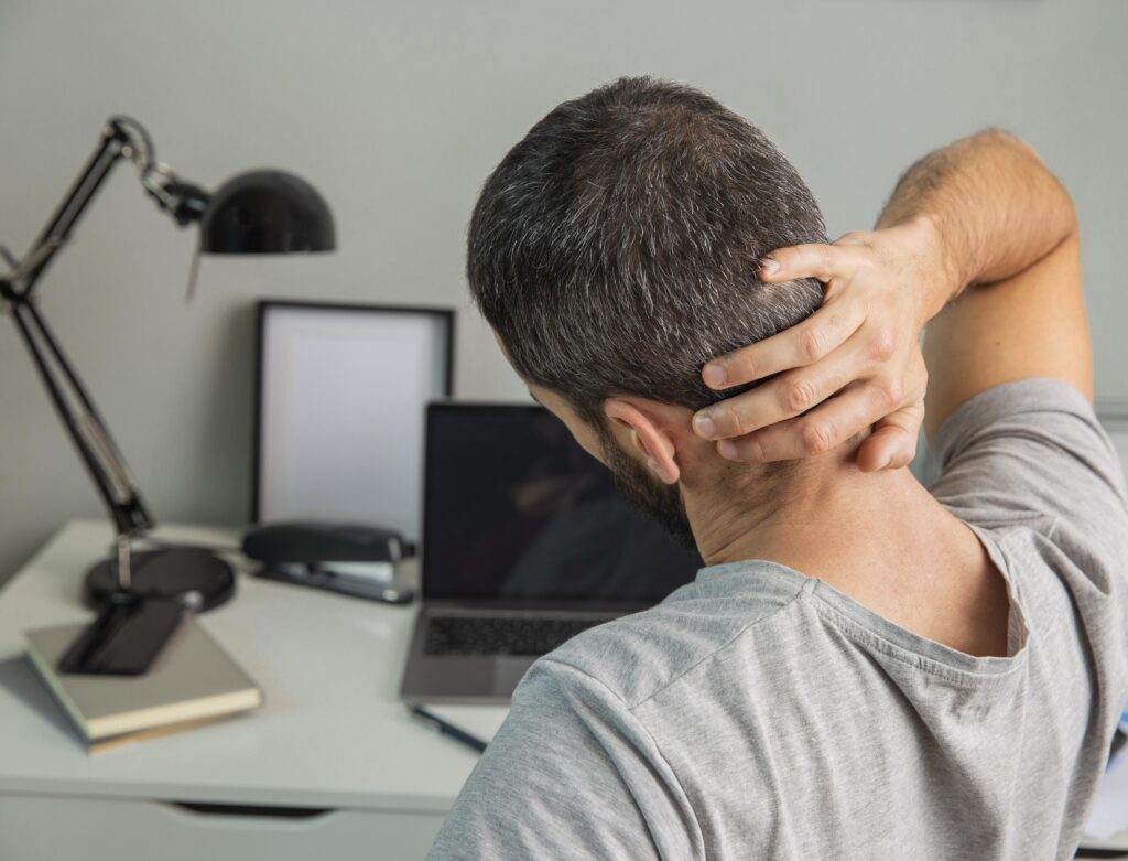 Imagine de aproape, din spate, bărbat care stă la laptop, acuză dureri de ceafă