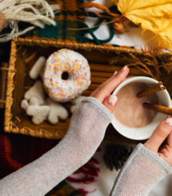 mainile unei femei cu o cana cu o bautura calda, cacao cu lapte, cu o gogoasa langa, decor de toamna