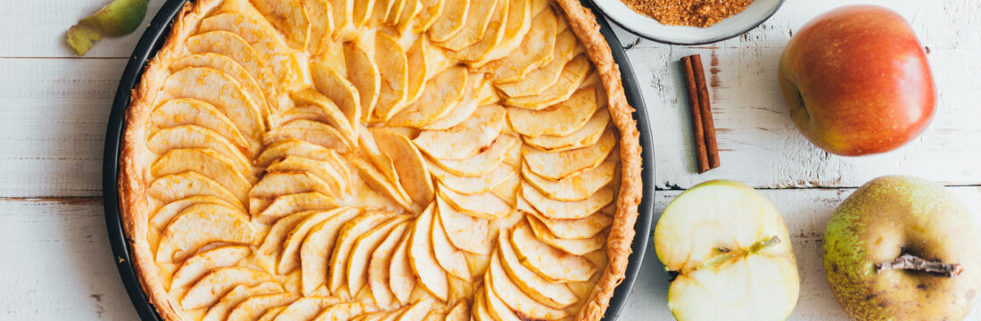 Imagine de sus, prajitura de mere de post pe un fundal de lemn alb. Lângă sunt felii de mere, bețișoare de scorțișoară și un bol cu scorțișoară rasă