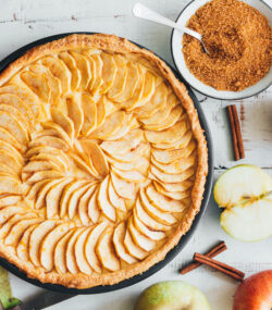 Imagine de sus, prajitura de mere de post pe un fundal de lemn alb. Lângă sunt felii de mere, bețișoare de scorțișoară și un bol cu scorțișoară rasă