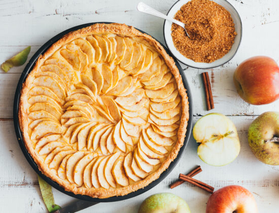 Imagine de sus, prajitura de mere de post pe un fundal de lemn alb. Lângă sunt felii de mere, bețișoare de scorțișoară și un bol cu scorțișoară rasă