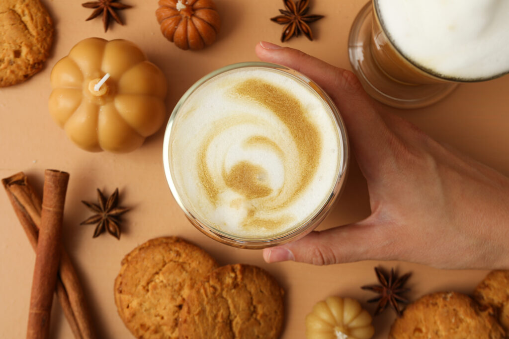 Imagine de sus, pahar cu pumpkin spice latte pe un fundal maro deschis, cu prăjituri, steluțe de anason și lumânări în formă de dovleac. O persoană ține paharul în mână