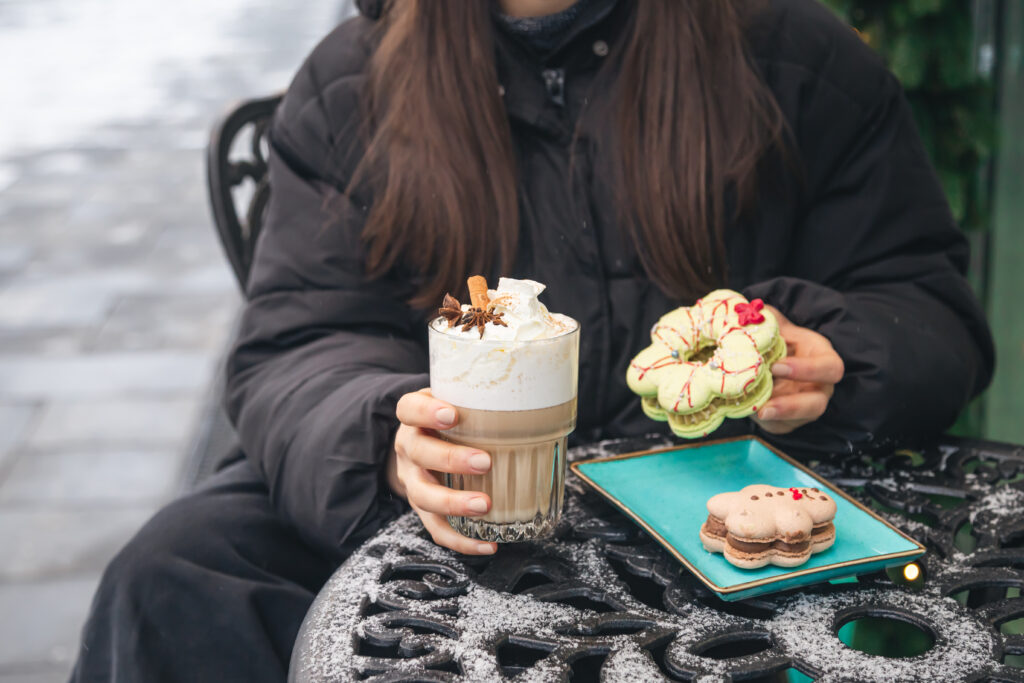 o femeie sta la masa afara iarna, pe timp de frig, tine in mana o prajitura cu crema si o cafea cu lapte