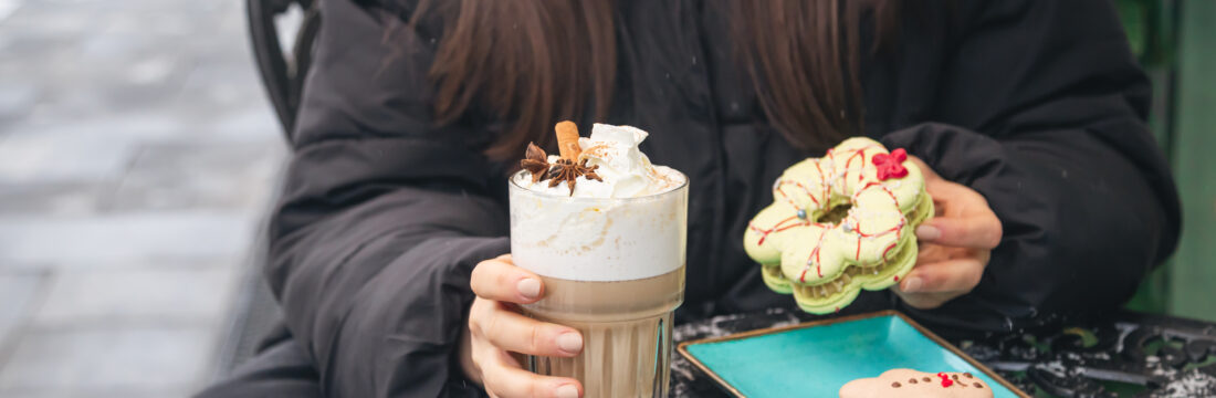 o femeie sta la masa afara iarna, pe timp de frig, tine in mana o prajitura cu crema si o cafea cu lapte