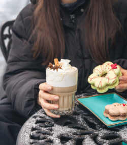 o femeie sta la masa afara iarna, pe timp de frig, tine in mana o prajitura cu crema si o cafea cu lapte