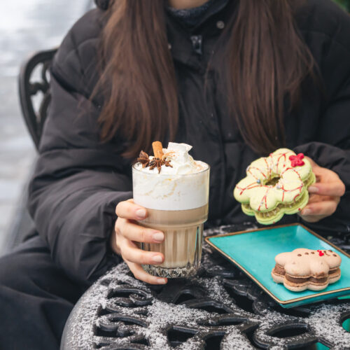o femeie sta la masa afara iarna, pe timp de frig, tine in mana o prajitura cu crema si o cafea cu lapte