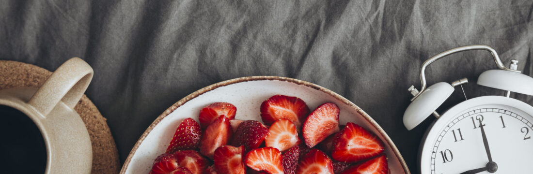 Imagine de sus, ceașcă de cafea, bol cu căpșuni și un ceas pe un cearceaf gri