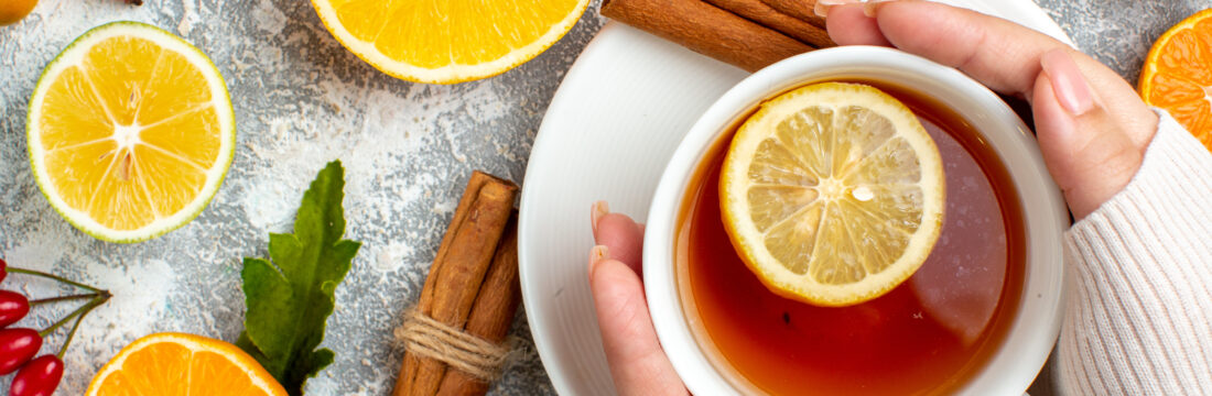 Imagine de sus, persoană care ține între palme o cană de ceai cu o felie de lămâie deasupra. Pe masă, alte felii de lămâie și bețișoare de scorțișoară