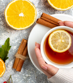 Imagine de sus, persoană care ține între palme o cană de ceai cu o felie de lămâie deasupra. Pe masă, alte felii de lămâie și bețișoare de scorțișoară