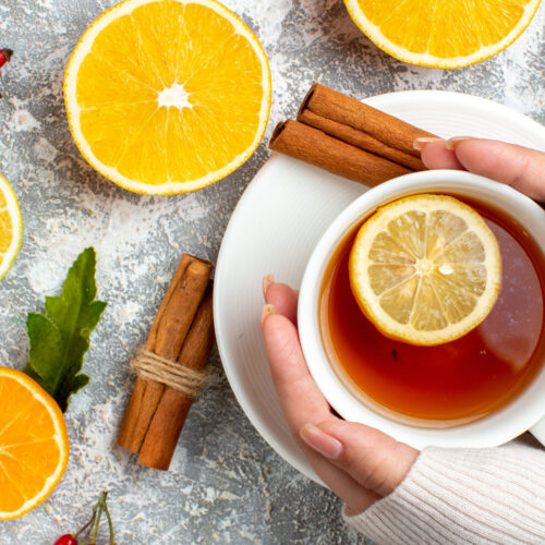Imagine de sus, persoană care ține între palme o cană de ceai cu o felie de lămâie deasupra. Pe masă, alte felii de lămâie și bețișoare de scorțișoară
