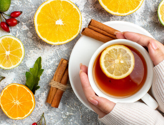Imagine de sus, persoană care ține între palme o cană de ceai cu o felie de lămâie deasupra. Pe masă, alte felii de lămâie și bețișoare de scorțișoară