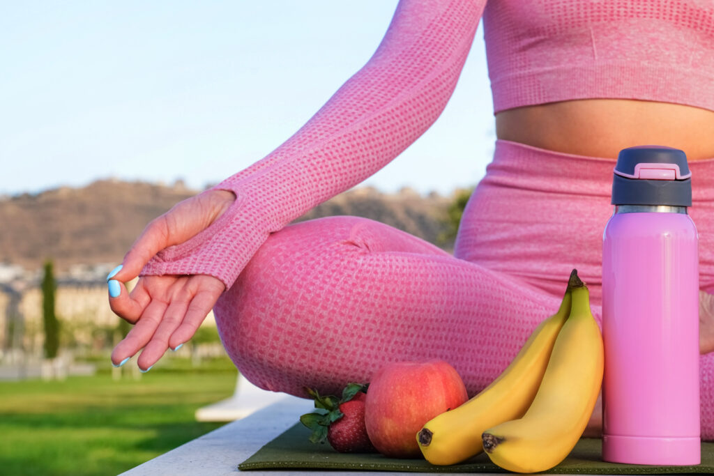 Imagine de aproape, persoană care stă în poziție de yoga. Lângă, are un termos de apă, 2 banane, un măr și câteva căpșuni