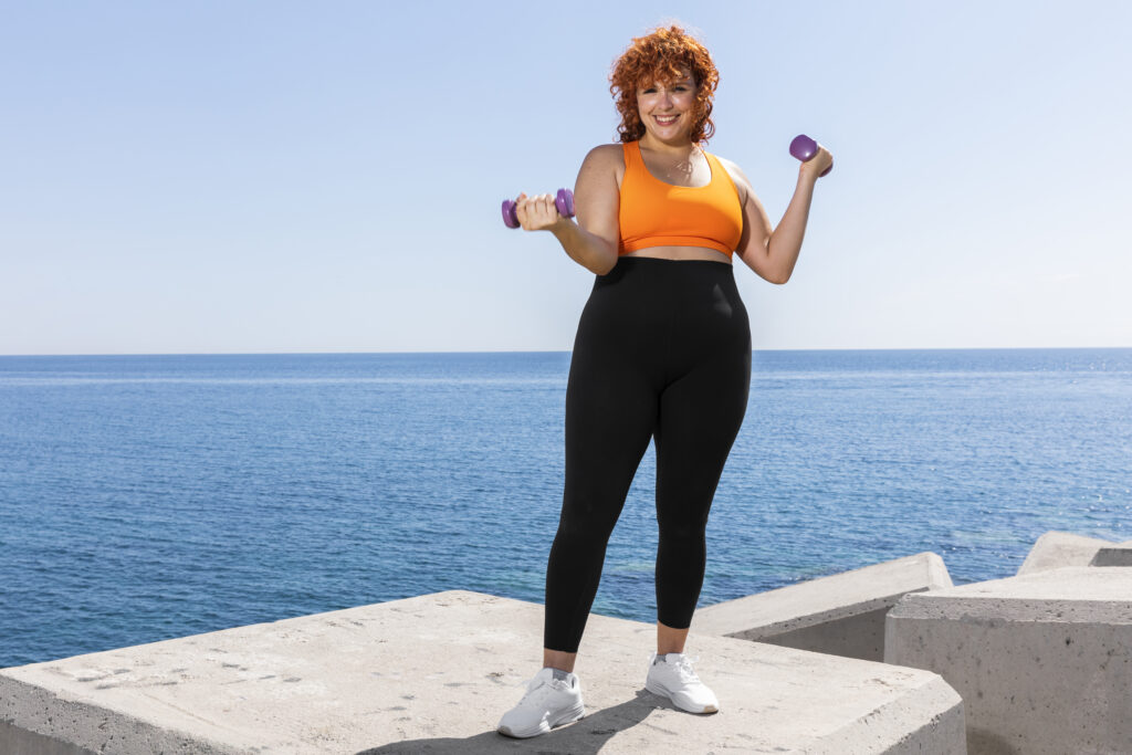 Femeie tânără îmbrăcată sport zâmbește către cameră în timp ce face exerciții cu greutăți pe faleză