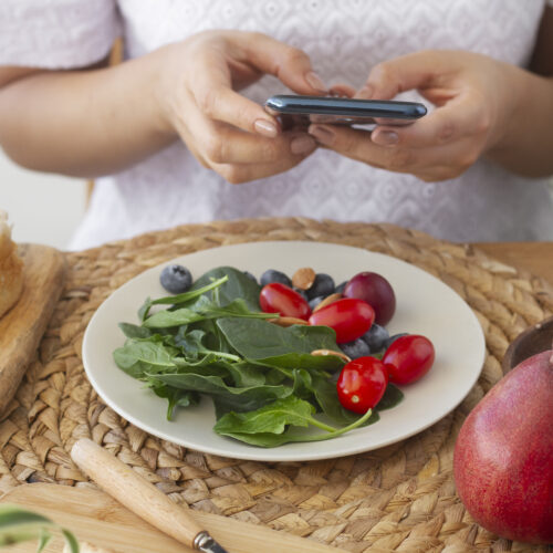 femeie sta la masa cu o salata sanatoasa in fata, tine in mana telefonul