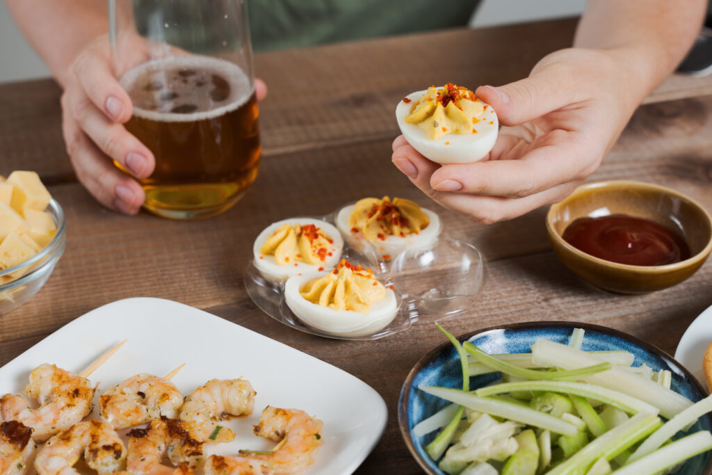 Imagine de aproape, persoană care ține într-o mână un ou umplut și în cealaltă un pahar de bere. Pe masă mai sunt creveți gătiți, ceapă verde și un sos roșu